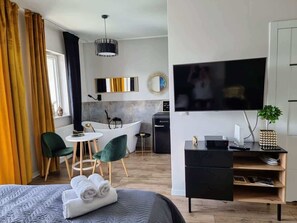A bedroom setup including a bed, a desk with a chair, a flat-screen TV mounted on the wall, and large curtains.