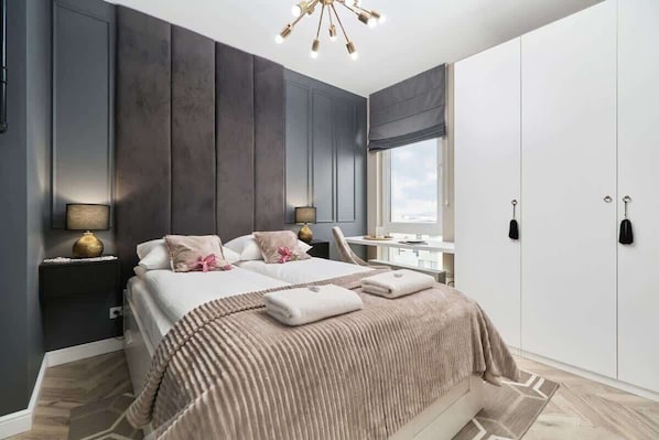 A bedroom with a plush double bed featuring beige and white bedding. A modern gray accent wall with soft lighting adds a luxurious touch.