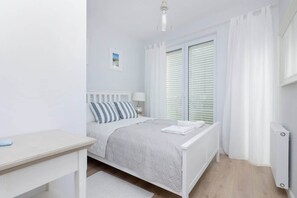 A cozy bedroom featuring a double bed, white linens, and bright lighting from a large window.