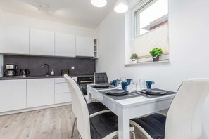 In the foreground is a white dining table with tableware. A kitchenette with white cabinetry, a sleek countertop, and minimalistic decor.