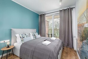 A cozy bedroom with a large bed, gray bedding, teal walls, and natural light from the windows.