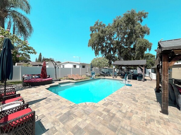 A stunning backyard oasis with a sparkling pool, surrounded by a spacious paved deck. Cozy lounge chairs, shaded pergolas, and lush greenery create the perfect retreat for relaxation and outdoor entertaining.