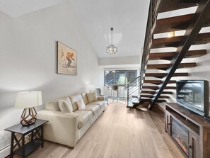 Modern living room with a beige couch, side table, and lamp on the left, a wall-mounted painting above, a staircase with dark wooden steps on the right, and a TV on a wooden unit underneath.