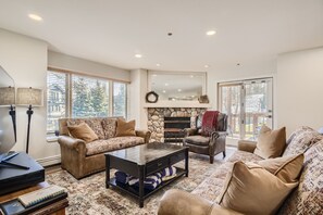 A cozy living room with a stone fireplace, two sofas, an armchair, a coffee table, and large windows letting in natural light. Decor includes a wreath above the fireplace and a throw blanket on the armchair.