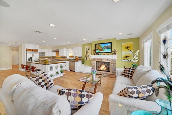 The main living area is located on the ground floor and includes two sofas, cable TV, fireplace, and kitchen and dining areas beyond.
                