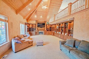 The spacious family room features fine woodwork. The three-story vaulted ceilings stretch high above, creating an airy expanse with wooden beams overhead. Built-in shelves flank the grand fireplace, and a Large Screen TV complete the room.