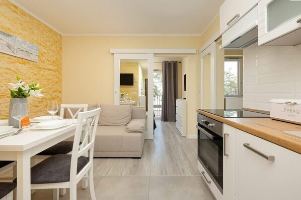 A bright living space with a yellow accent wall, a dining table with white chairs, and an open-plan kitchen in the background.