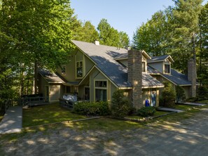 Two-story house with beige siding and stone chimney, surrounded by trees. Front door with two steps and a small front yard. Driveway leading up to the house on a sunny day.