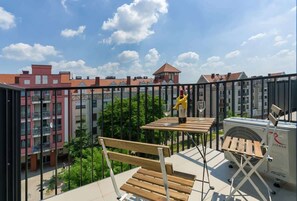 A cozy balcony featuring a table with chairs, offering a view of surrounding red-roofed buildings and greenery under a bright sky.