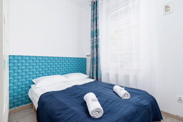 A cozy bedroom with a blue headboard, white bedding, and neatly folded towels on the bed. A small lamp is visible on the bedside table.