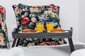 A floral tray with breakfast items, including a glass of orange juice, fruit, and a croissant, set on a sofa with decorative cushions.