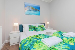 A bedroom featuring a bed with vibrant green, tropical-themed bedding and matching pillows. A small bedside table with a lamp is visible.