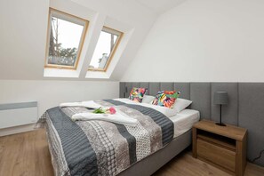 A cozy bedroom with slanted ceilings and skylight windows, featuring two single beds, a colorful blanket, and storage space. The room has a cozy cabin-like feel due to the wooden finishes.