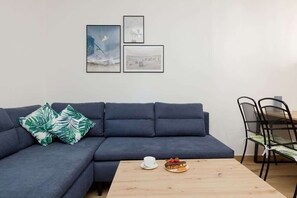 A spacious living room featuring a dark navy-blue sectional sofa, a wooden coffee table, and minimalistic decor with soft lighting and a small dining area in the background.