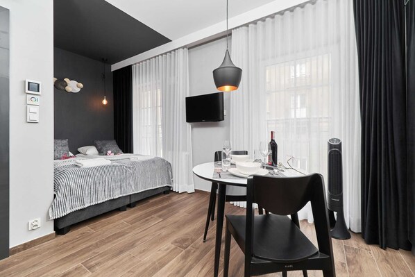 A modern studio apartment featuring a living room with a double bed, a kitchenette in the background, and light wooden flooring. The design is minimalist and functional.