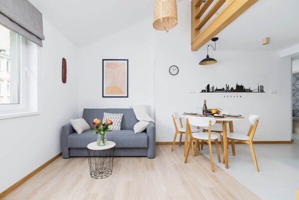A bright living room featuring a grey sofa, a small round coffee table, and a dining table with wooden chairs under a slanted ceiling.