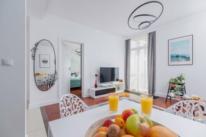 In the foreground, a white dining table with white chairs and a fruit bowl lying on the table is visible. On the other side There is a flat-screen TV.