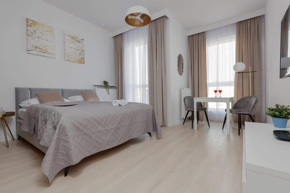 A spacious, modern studio with soft beige tones, featuring a large bed, a seating area with a table, and ample natural light coming through large windows.