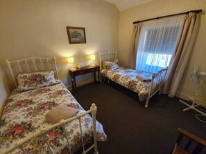 This charming twin bedroom features two vintage-style iron beds adorned with floral bedding, creating a cozy and nostalgic ambiance. A wooden nightstand with soft lighting sits between the beds, while a large window with sheer curtains allows gentle natur