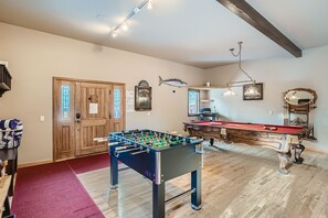 A game room featuring a foosball table, a pool table, a dartboard, and a wall-mounted fish decoration. The room has wooden flooring, light-colored walls, and a wooden door.