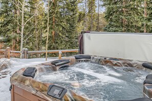 A hot tub with bubbling water is placed outdoors on a snow-covered deck in front of a forested area with tall evergreen trees.