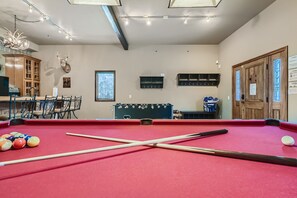 Room with a red felt pool table, two pool cues on the table, a foosball table, bar area with stools, wall-mounted shelves, and a wooden entry door. Decor includes lights and a mounted animal head.