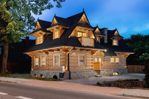 A traditional wooden chalet surrounded by greenery, set against a backdrop of mountains. The architecture highlights charming rustic details.