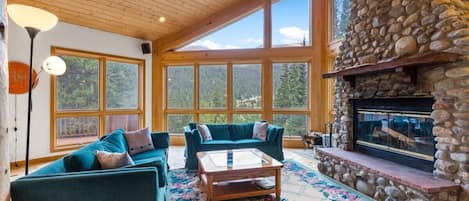 Relax in the spacious family room with floor-to-ceiling windows,  a cozy indoor wood fireplace, flat-screen TV, piano, and sofa with seating for up to 5 people