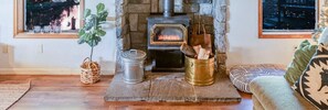 A charming corner with a burning stove set against a stone fireplace offers a tranquil retreat, enveloped by the natural light filtering through the windows.