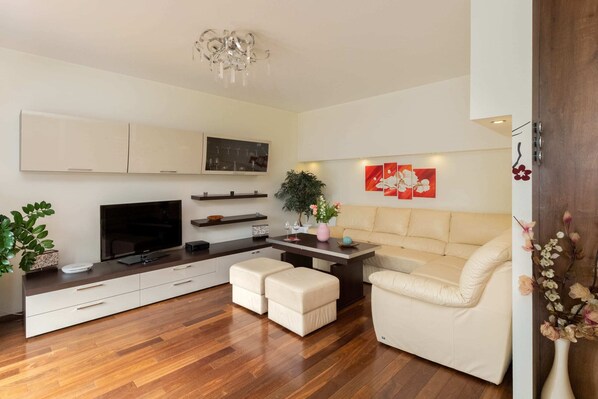 A bright living room with wooden flooring, white sofas, a TV unit, and stylish decorative plants, offering a cozy and inviting atmosphere.