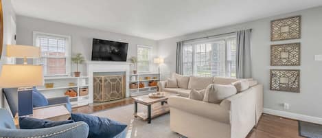 Spacious and stylish living room in our Airbnb with seating for 10 and a large, wall-mounted smart TV. Modern decor, neutral color palette, and ample natural light create a calming atmosphere. Perfect for relaxing after a day of exploring and spending time with loved ones.