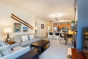 A modern living room with a gray sofa, wooden coffee table, wall-mounted skis, and a kitchen area in the background featuring light wood cabinets and stainless steel appliances.