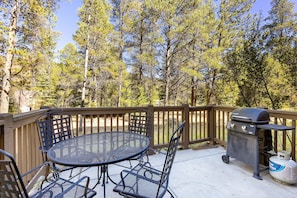 Outdoor deck with a black metal table and four chairs set, a gas grill, and surrounded by tall pine trees.