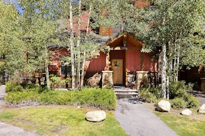 A red wooden house with a stone foundation and covered porch, surrounded by trees and shrubs, viewed from the front. The house is located in a wooded area with a concrete path leading to the entrance.