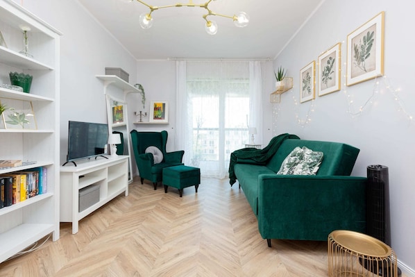 A stylish living room featuring a green velvet sofa, white shelves, and golden accents, illuminated by natural light from large windows.