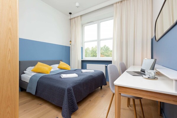 A bright bedroom with a double bed, blue and yellow accents, and natural light streaming through the window.
