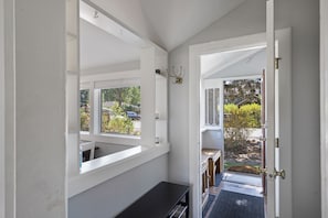 View of a small entryway with an open door leading outside. The space features a coat hook, bench, and large windows. Light pours in from the outside, creating a bright and welcoming atmosphere.