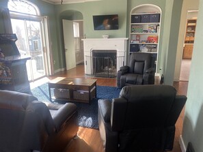 Living Room showing Roku TV with DVD player, fireplace, shelves with board games, sofa sleeper and coffee table, a part of the dining room and kitchen are partially shown to the right