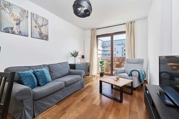 A modern living room with a gray sofa, wooden flooring, a glass coffee table, and a large window letting in natural light.