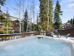 Outdoor hot tub on a snowy deck, surrounded by tall trees and adjacent buildings in a forested area.