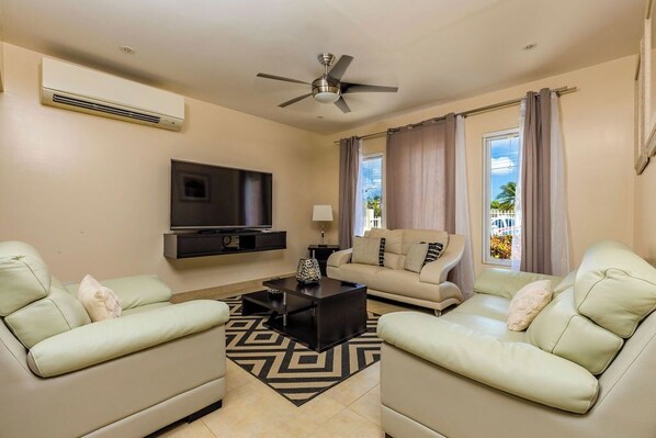 A living room filled with a comfortable sofa. There is an air conditioning unit and ceiling fan. There's a TV for entertainment and large curtains for privacy