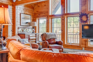 A cozy, sunlit living room with wooden walls, a large orange couch, an armchair, and a decorative wreath above the windows. A landscape painting and a lamp are placed on a wooden dresser.