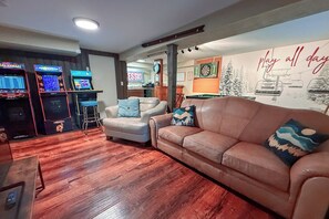 The open living and game area downstairs with 3 arcade games, flat screen tv, bumper pool, darts, Hook 21, and shot ski (21+). It is connected to kitchenette area. 