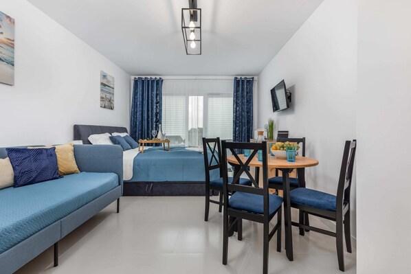A living room with a blue sofa and a dining area in the background, featuring a simple table and chairs, all styled in modern, neutral tones.