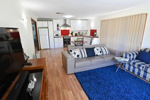 This open-concept living area combines a cozy lounge and a stylish kitchen. It features a grey leather sofa with patterned cushions, a checkered armchair, and a vibrant blue rug. The kitchen in the background includes modern white cabinetry, red accents, 
