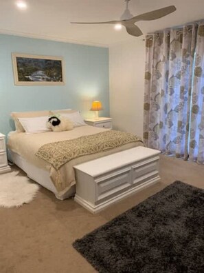 This cozy bedroom features a queen-sized bed with neutral bedding, flanked by matching white nightstands and a chest at the foot. A soft blue accent wall adds a calming touch, complemented by floral curtains and plush rugs. A ceiling fan and warm lighting