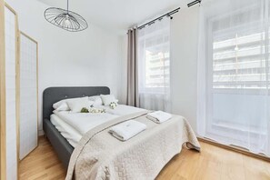 A bright bedroom with a double bed, white bedding, and a large window for natural light.
