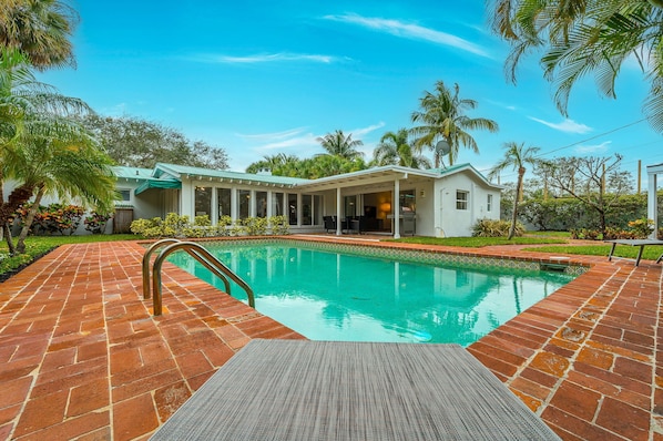 Resort-style heated pool with sun loungers, outdoor wicker seating and dining, and amazing landscaping with tropical palms, plants, and flowers.