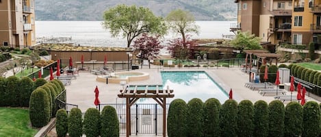 Relax in style at this resort-like pool area, featuring a serene lake view, red umbrellas, and cozy lounge chairs. A wooden pergola offers shade, while rolling mountains set a peaceful backdrop.
