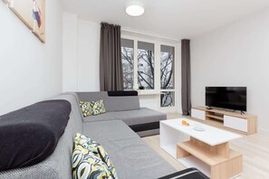 This sleek living room offers a blend of elegance and comfort, showcasing a TV, spacious seating, and large windows that let in abundant natural light. Ideal for a cozy family evening or home cinema experience.